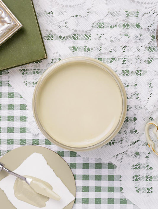 A jar filled with pale green paint sits on a lace and green checkered tablecloth, surrounded by a palette knife, a green book, and part of a teacup.