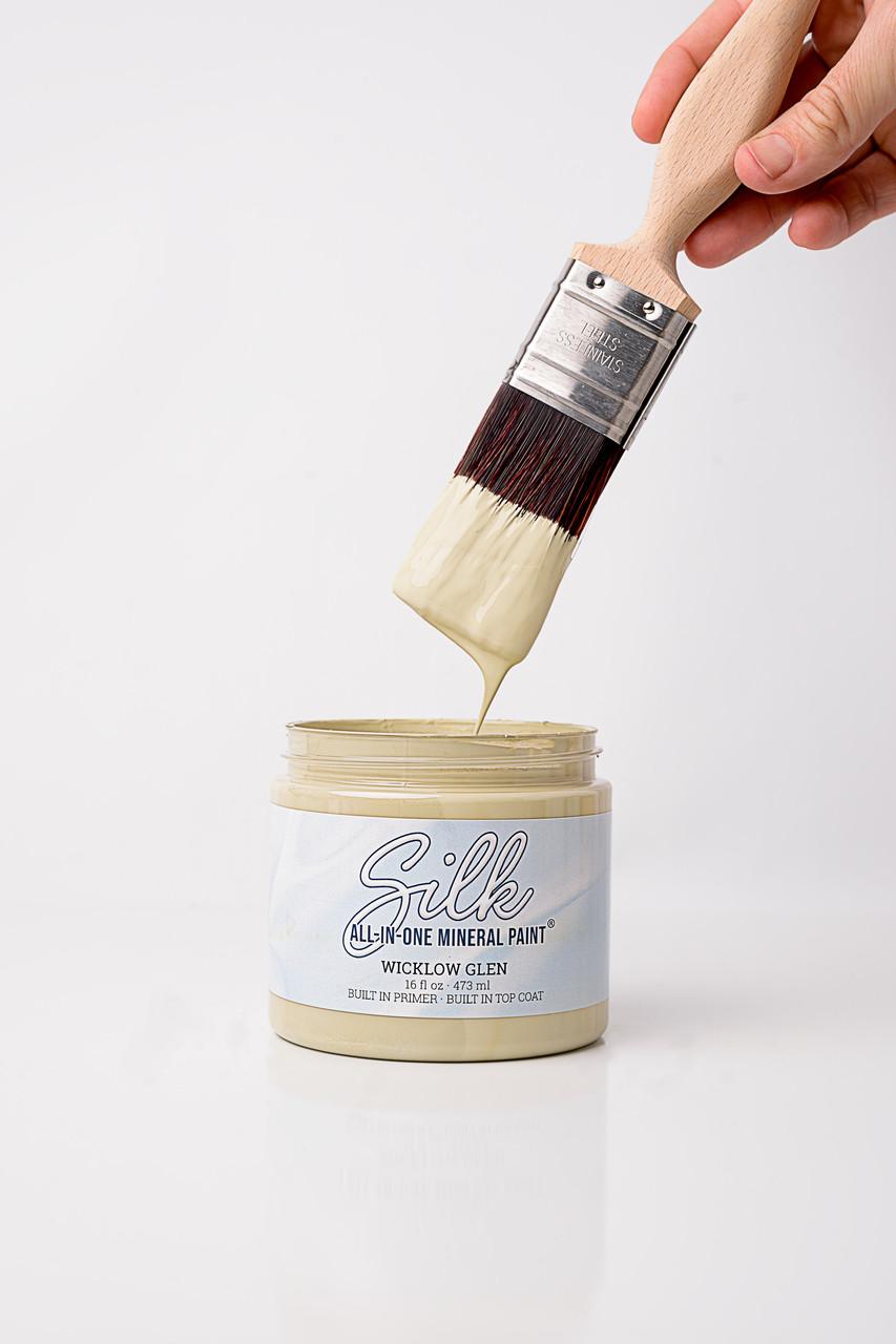 A hand holds a paintbrush dipped in light green paint above an open jar labeled "Silk All-In-One Mineral Paint, Wicklow Glen." Paint is dripping from the brush into the jar against a white background.