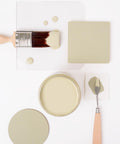 A paintbrush with light green paint, a jar of matching paint, a square sample, and a palette knife with paint are arranged neatly on a white surface.