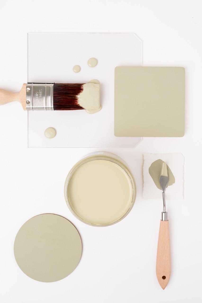 A paintbrush with light green paint, a jar of matching paint, a square sample, and a palette knife with paint are arranged neatly on a white surface.