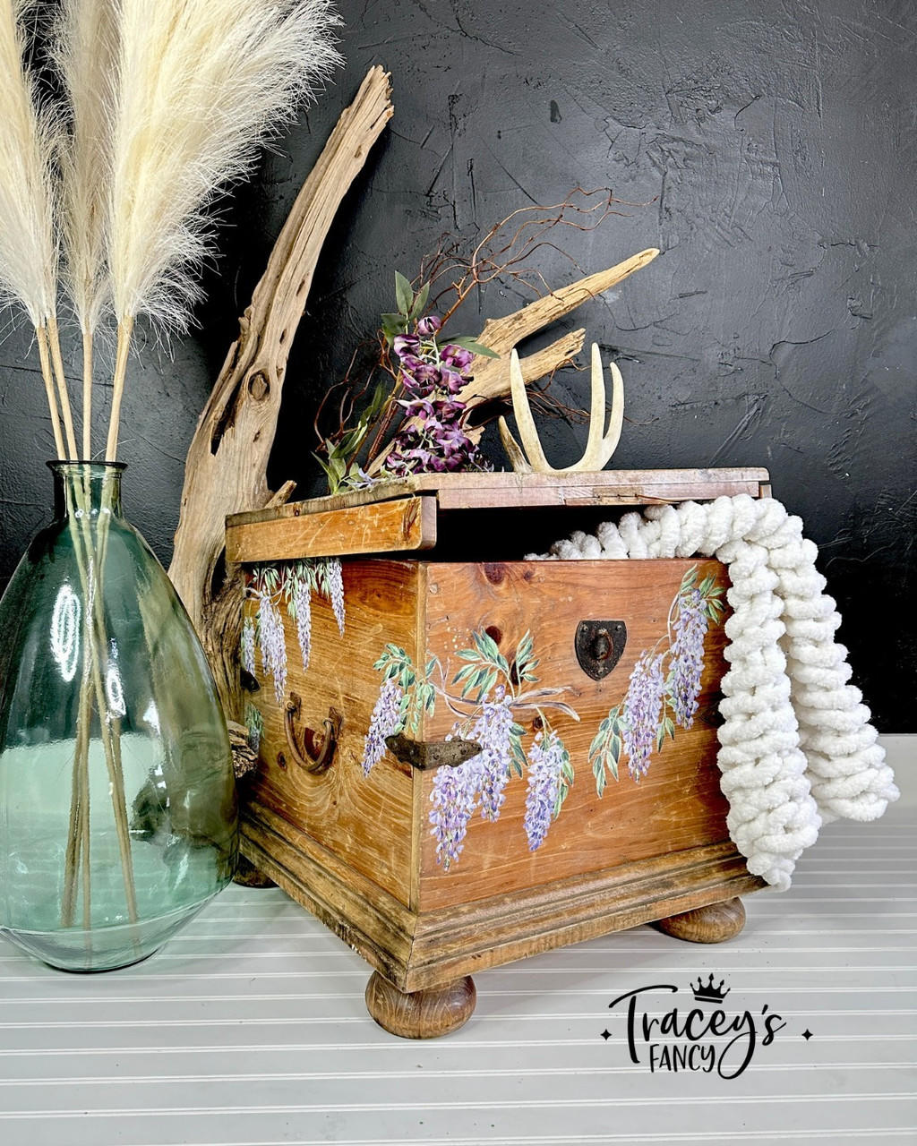 A natural wood pine box with white macrame yarn flowing out of it. The Wisteria Spring Transfer has been applied to the front and side. Staged with driftwood behind it with purple flowers and a clear vase holding feathers beside it.