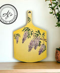 A yellow wooden tray with handle. You can see Wisteria Spring Transfer on the front of the wooden board and on the handle. Staged next to a plant inside a woven pot.