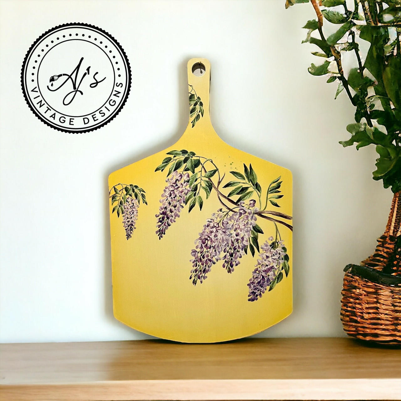 A yellow wooden tray with handle. You can see Wisteria Spring Transfer on the front of the wooden board and on the handle. Staged next to a plant inside a woven pot.