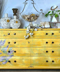 A yellow painted dresser with bronze hardware, you can see the Wisteria Spring Transfer applied to the front of the drawers. Staged with white circular vases with flowers on them on top, next to a tall wire baskets holding rocks, next to a stack of gold books with twine tied around them. White faux flowers on top of the dresser hanging down.