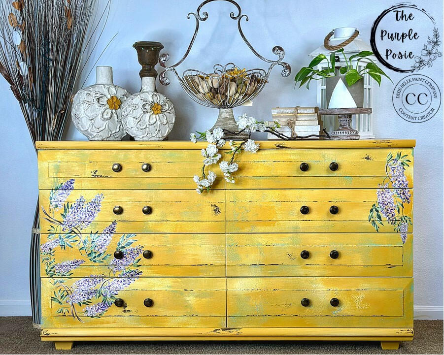 A yellow painted dresser with bronze hardware, you can see the Wisteria Spring Transfer applied to the front of the drawers. Staged with white circular vases with flowers on them on top, next to a tall wire baskets holding rocks, next to a stack of gold books with twine tied around them. White faux flowers on top of the dresser hanging down.