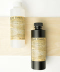 Two bottles labeled "Wood Bleach" are placed on a light wooden board. One bottle is clear with a white cap, and the other is black with a black cap. Both have beige labels with detailed text.