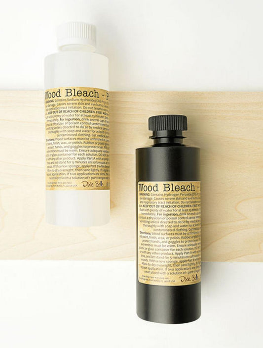 Two bottles labeled "Wood Bleach" are placed on a light wooden board. One bottle is clear with a white cap, and the other is black with a black cap. Both have beige labels with detailed text.