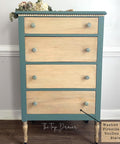 A four-drawer dresser with natural wood drawer fronts, teal blue sides and top, beaded trim, floral knobs, and decorative flowers placed on top, standing against a pale wall on a wooden floor.