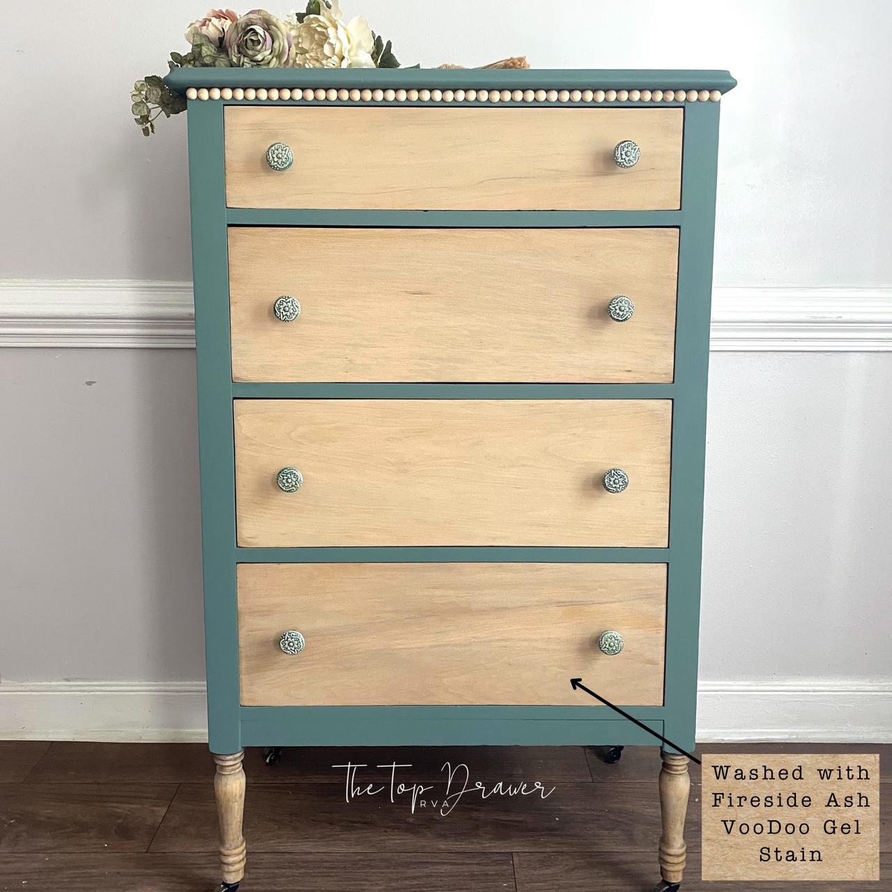 A four-drawer dresser with natural wood drawer fronts, teal blue sides and top, beaded trim, floral knobs, and decorative flowers placed on top, standing against a pale wall on a wooden floor.