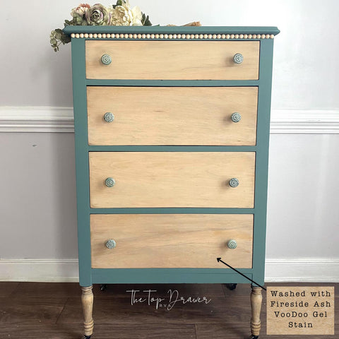 A four-drawer dresser with natural wood drawer fronts, teal blue sides and top, beaded trim, floral knobs, and decorative flowers placed on top, standing against a pale wall on a wooden floor.
