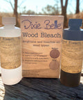 Two bottles and a box of Dixie Belle Wood Bleach are displayed on a wooden surface outdoors. The box highlights that the product brightens and neutralizes orange tones on wood. Sunlight shines in the background.