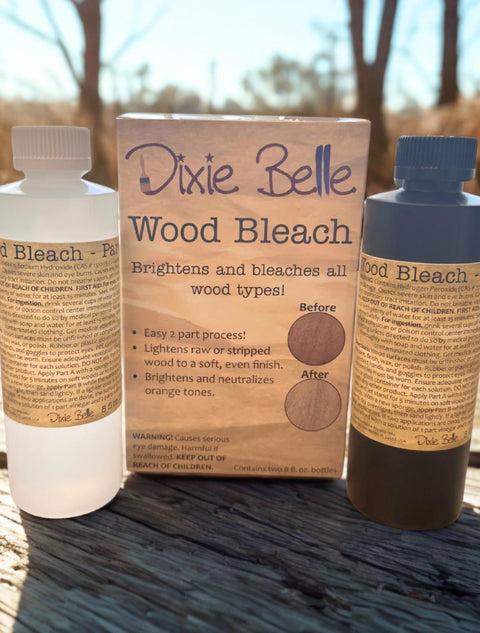 Two bottles and a box of Dixie Belle Wood Bleach are displayed on a wooden surface outdoors. The box highlights that the product brightens and neutralizes orange tones on wood. Sunlight shines in the background.