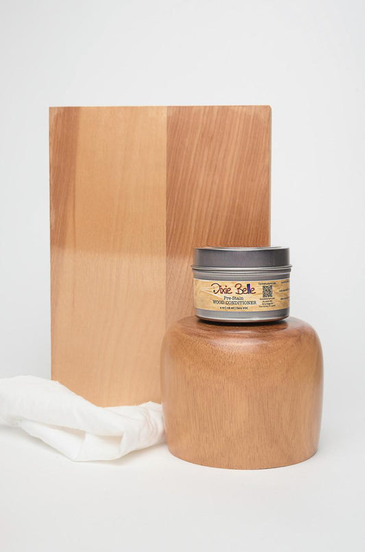A jar of Dixie Belle wood conditioner is placed in front of a wooden block and a polished wooden object. A white cloth rests beside the jar, all set against a plain white background.
