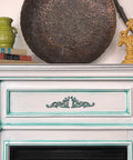 WoodUBend Applique #1361 (Set of 2) is the perfect mouldable embellishment for any project! This image shows Applique #1361 mounted on each side of a fireplace painted with Dixie Belle Chalk Mineral Paint. The fireplace is white with green accents. On the mantle are books, a vase and a metal tray.