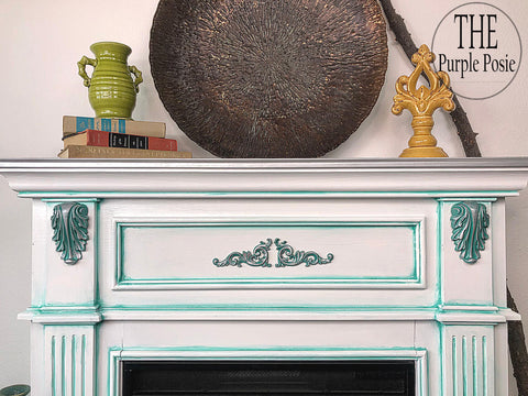 WoodUBend Applique #1361 (Set of 2) is the perfect mouldable embellishment for any project! This image shows Applique #1361 mounted on each side of a fireplace painted with Dixie Belle Chalk Mineral Paint. The fireplace is white with green accents. On the mantle are books, a vase and a metal tray.