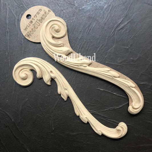 WoodUBend Applique #1723 (set of 2) is the perfect mouldable embellishment for any project!
The Applique #1723 is seen staged on top of a black crinkled background. One of the applique is seen in the packaging while the other is out.