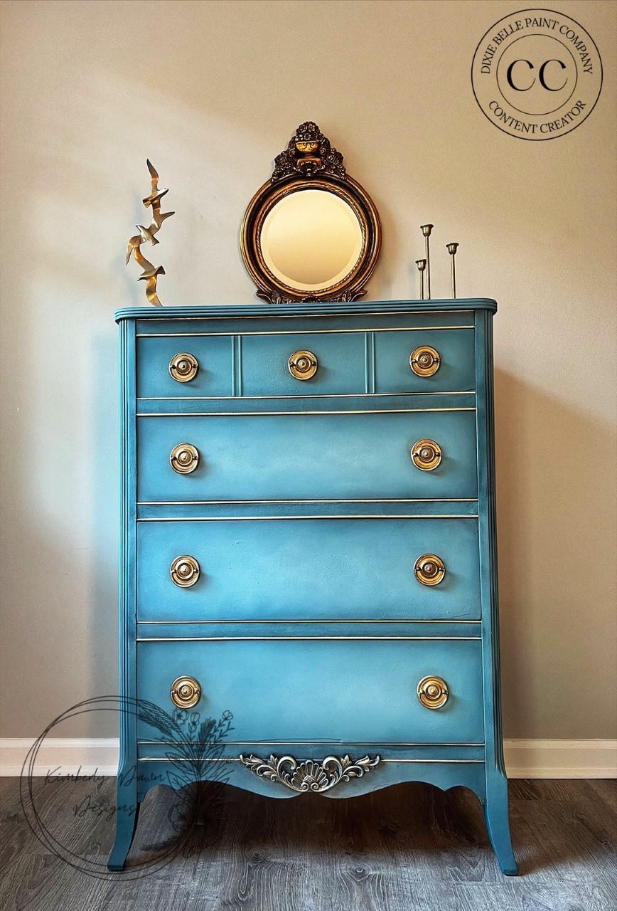 Applique #2152 Set of 2 is the perfect mouldable embellishment for any project!
A blue chest of drawers with gold hardware and at the bottom trim is seen Applique #2152 painted in silver. Staged on top of the piece is a bronze framed mirror.