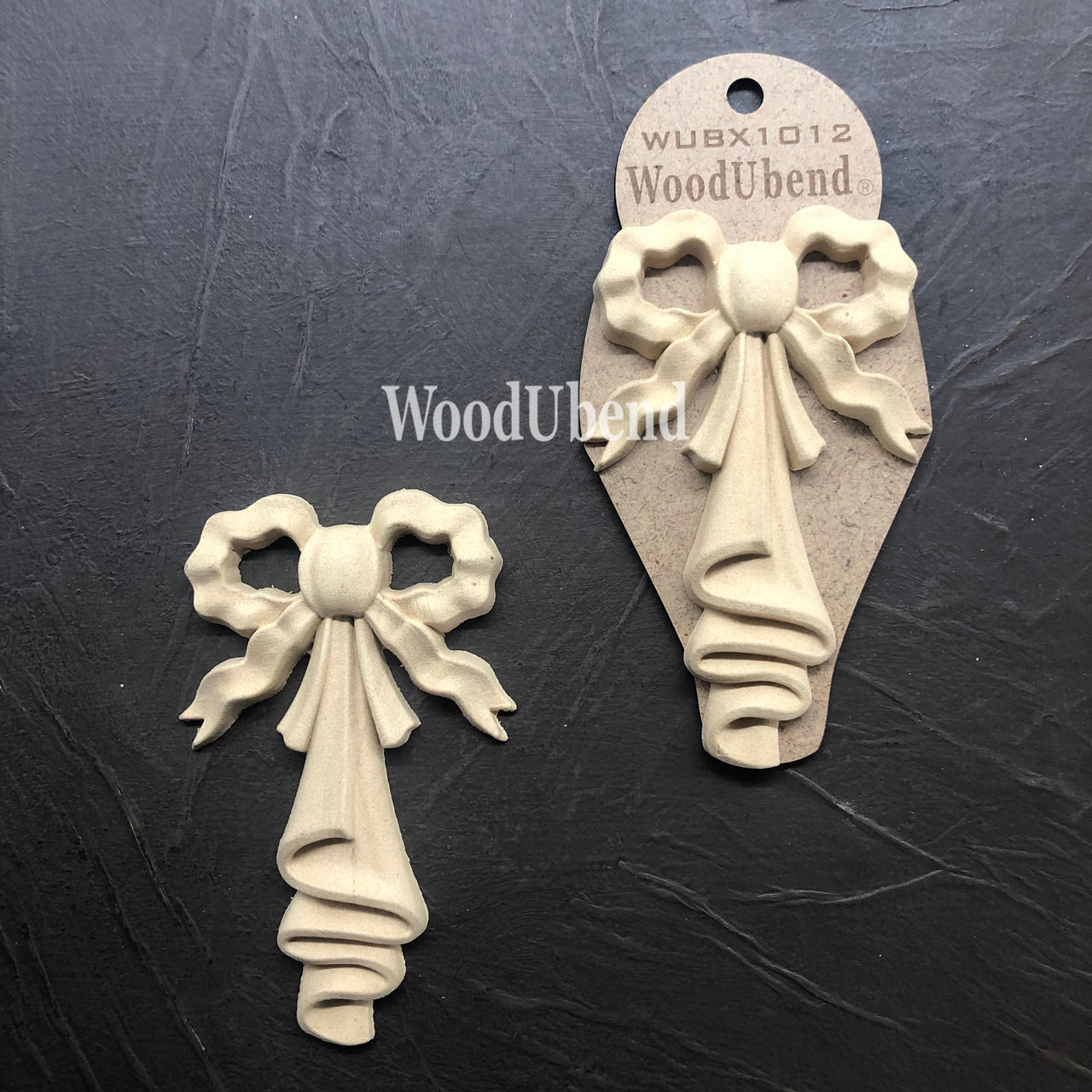 WoodUBend Bow Applique #X1012 (set of 2) is the perfect mouldable embellishment for any project!
This is a set of Wood-u-Bend bows.  The bows look wavy and there's a folded "linen" hanging down from the center of the bow.  The entire piece is a beige color.  One is out of the packaging and the other is still in the packaging.  They are both on top of crinkled,, black leather.