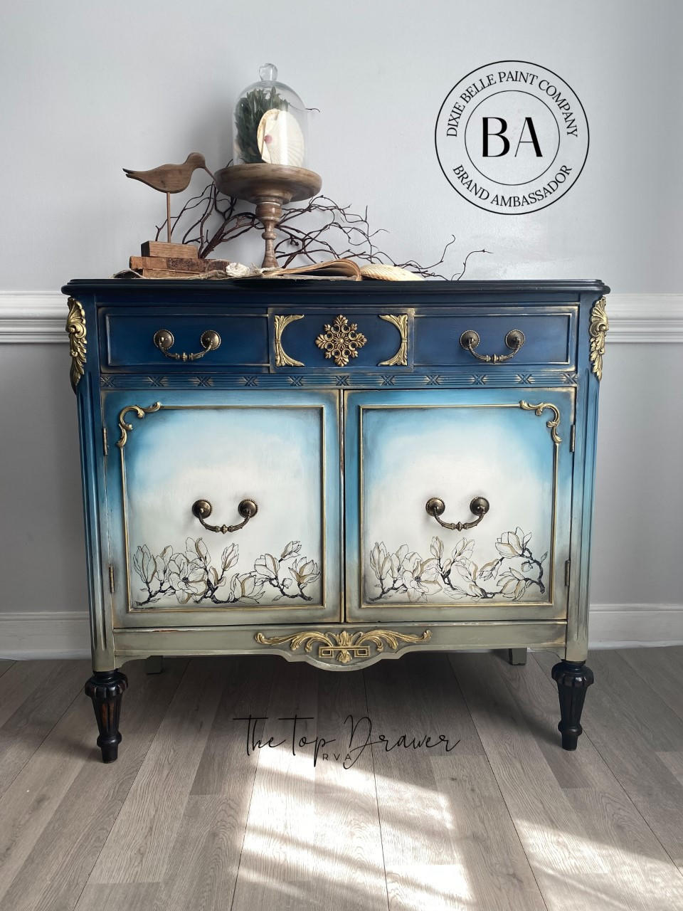 WoodUBend Floral Applique #2427 (set of 2) is the perfect mouldable embellishment for any project!
A credenza with the top of piece a dark blue going down to a mute green  with the middle front panels a blended white with Buds & Branches Transfer at the bottom. The  Floral Applique #2427 is seen on the top middle panel in-between two drawers painted gold.