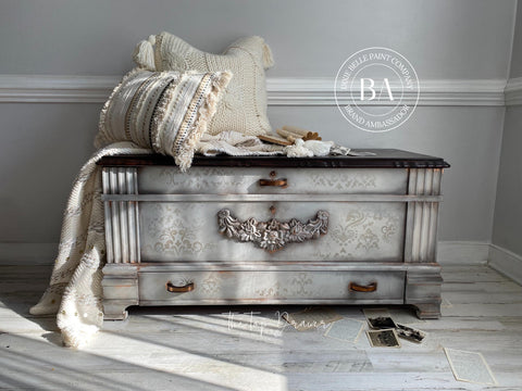 WoodUBend Floral Applique #X1001 is the perfect moldable embellishment for any project!
A chest with the middle front panel is The Floral Applique #X1001 in gray, with a bronze handle. Staged with white pillow cushions and a white blanket on top of the chest.