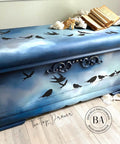 WoodUBend Ring Applique #1520 (set of 2) A trunk painted in white, blue, and black and blended as the night sky featuring the moulds and birds on a line in the front and on top. This trunk is styled with a blanket and book on the right side.