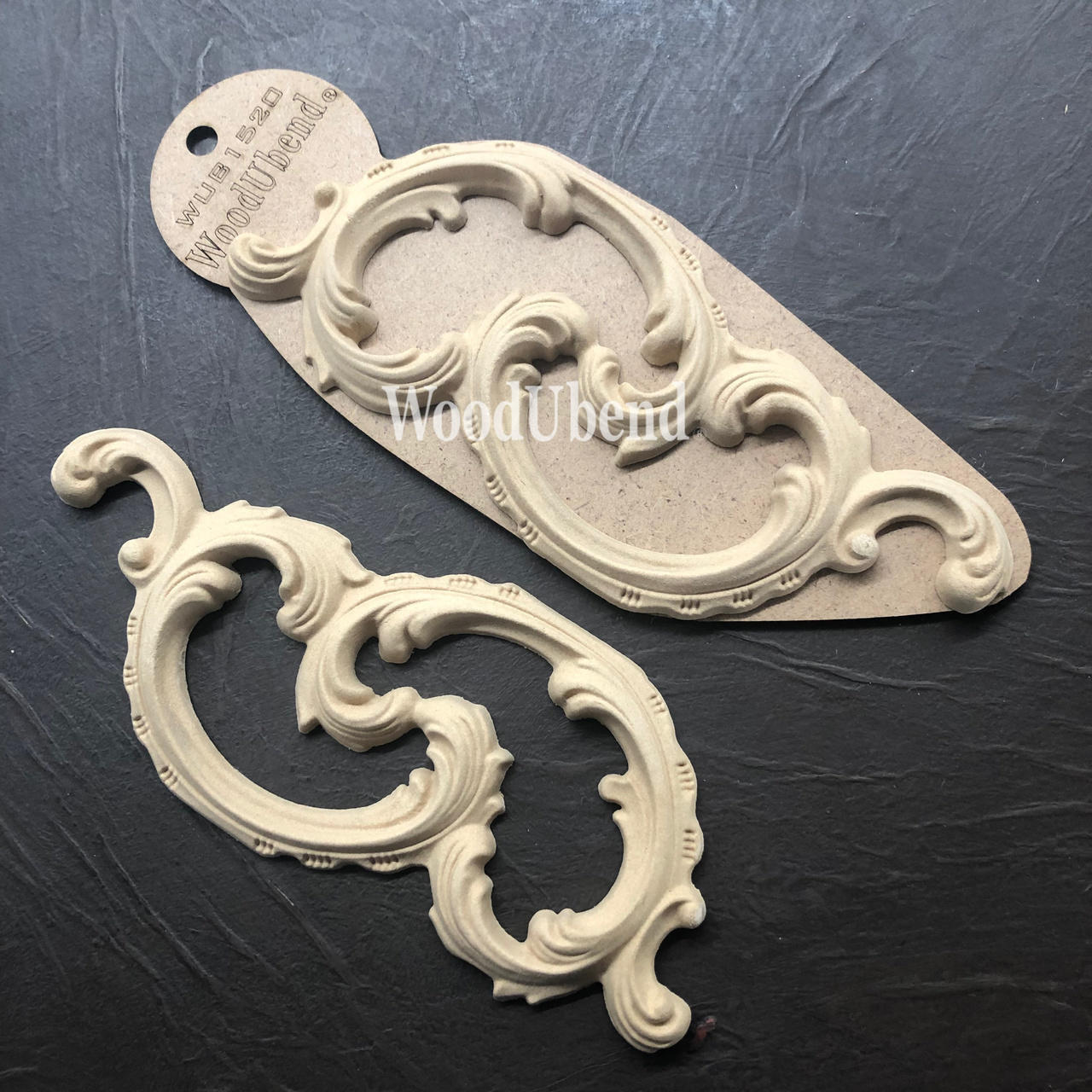 WoodUBend Ring Applique #1520 (set of 2) is the perfect mouldable embellishment for any project! A scroll style piece of wood applique placed beside each other angled to the left. The left applique is shown out of the package, and the right one is shown in the package.