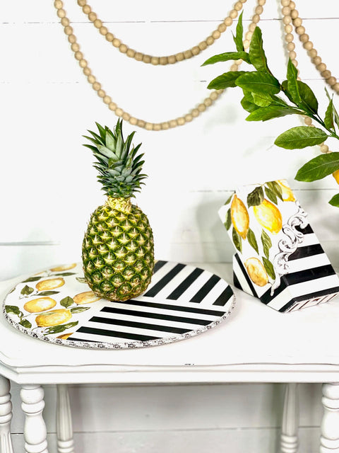 WoodUBend Trim #TR50a is the perfect mouldable embellishment for any project! A round tray and knife block that have black and white chalk painted stripes and lemons & leaves rub-on transfers.  WoodUBend Trim #TR50a is around the edge of the round tray, and a different WoodUBend moulding is on the knife block.  Both pieces are sitting on a white chalk painted table, with a pineapple on top of the tray.  In the background is greenery and strings of wooden beads.