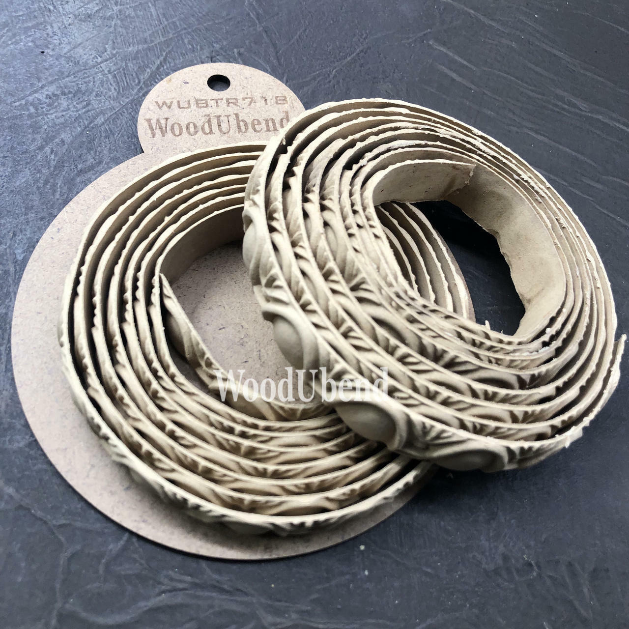 WoodUBend Trim #TR718 (Set of 2) is the perfect mouldable embellishment for any project!  Two rolls of WoodUbend sitting on top of a brown round cardboard cutout with WoodUbend WUBTR718 printed at the top; all sitting on a black table.