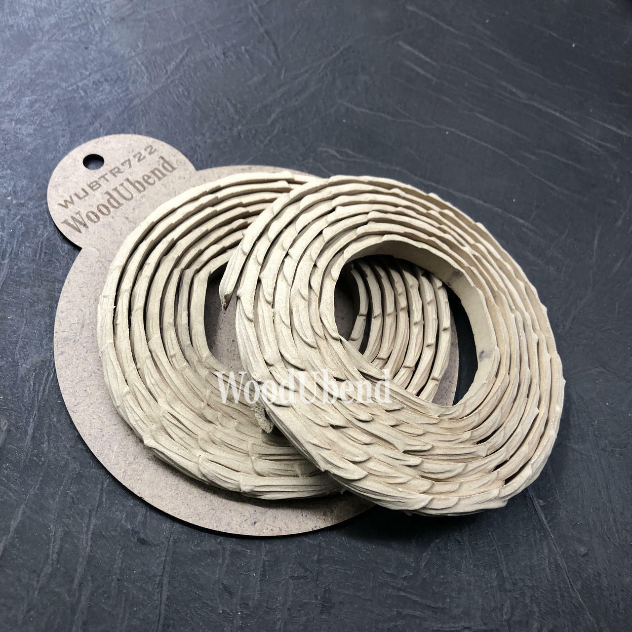 WoodUBend Trim #TR722 (Set of 2) is the perfect mouldable embellishment for any project!  Two rolls of WoodUbend sitting on top of a brown round cardboard cutout with WoodUbend WUBTR722 printed at the top; all sitting on a black table.