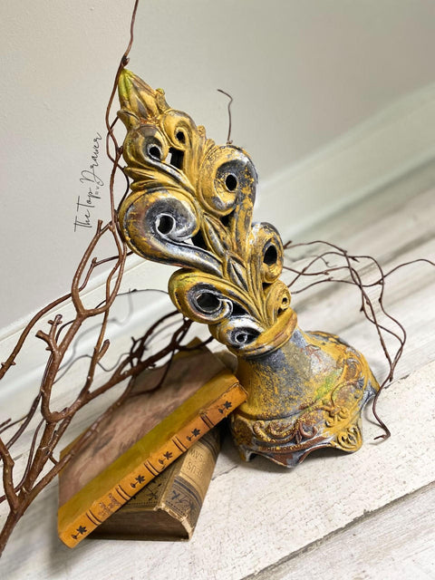 Fleur- de-lis type statue sprayed with Yellow Patina Spray. Staged with stacked books and a branch.