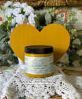 Yellow painted wooden heart with a jar of Yellowstone in front of it. Staged on crocheted lace with faux flowers and greenery in the background.